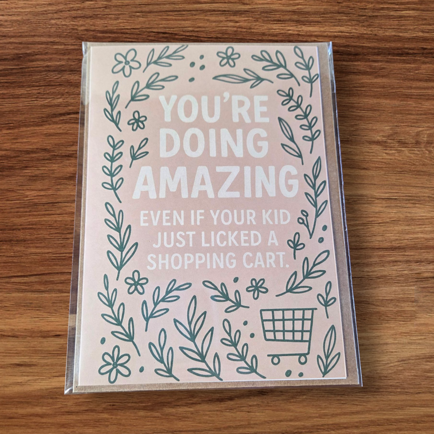 Greeting card reading ‘You’re Doing Amazing Even If Your Kid Just Licked a Shopping Cart’ with green floral border on blush background.