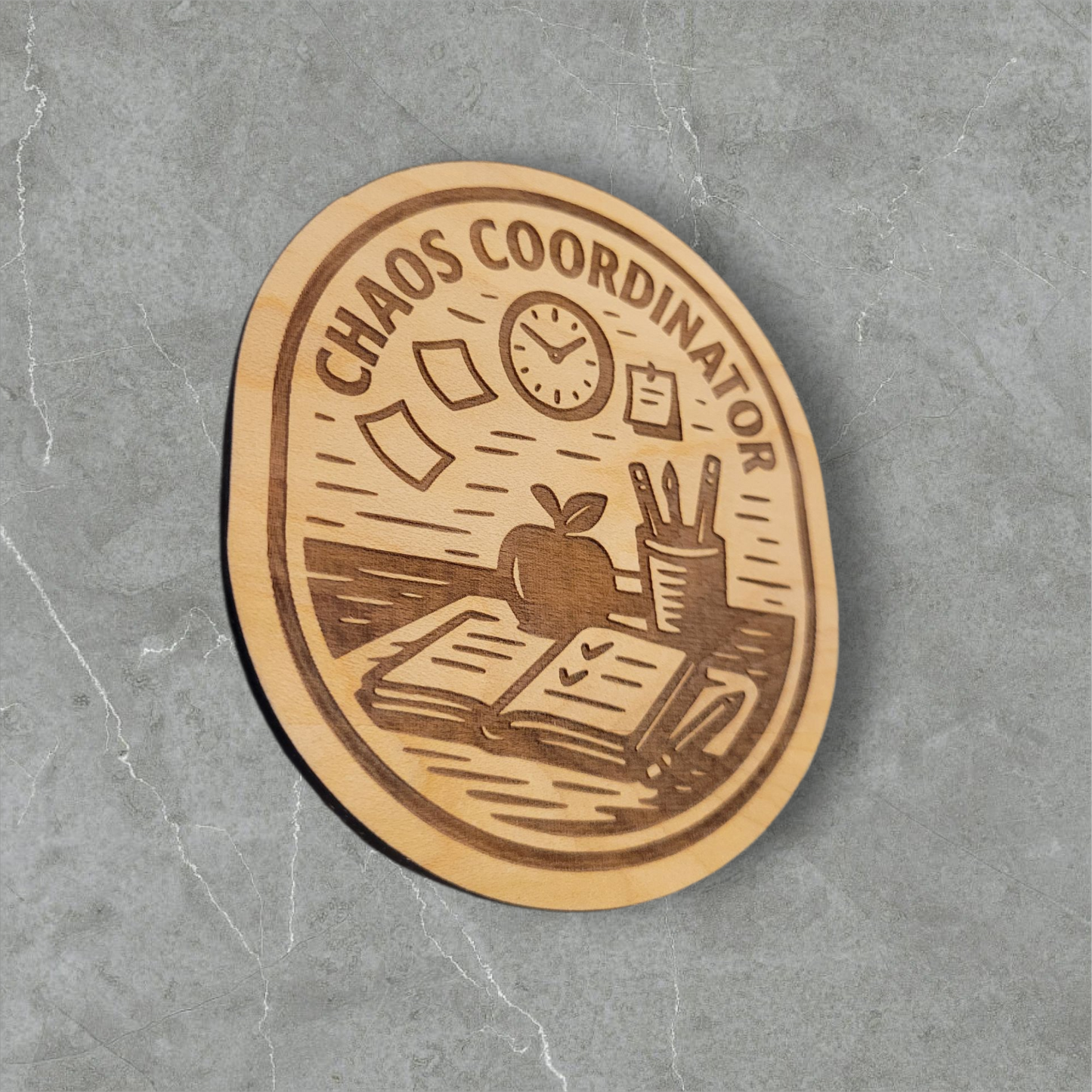 Laser engraved Chaos Coordinator wood magnet featuring teacher desk scene with open planner, apple, pencils, sticky notes, and clock