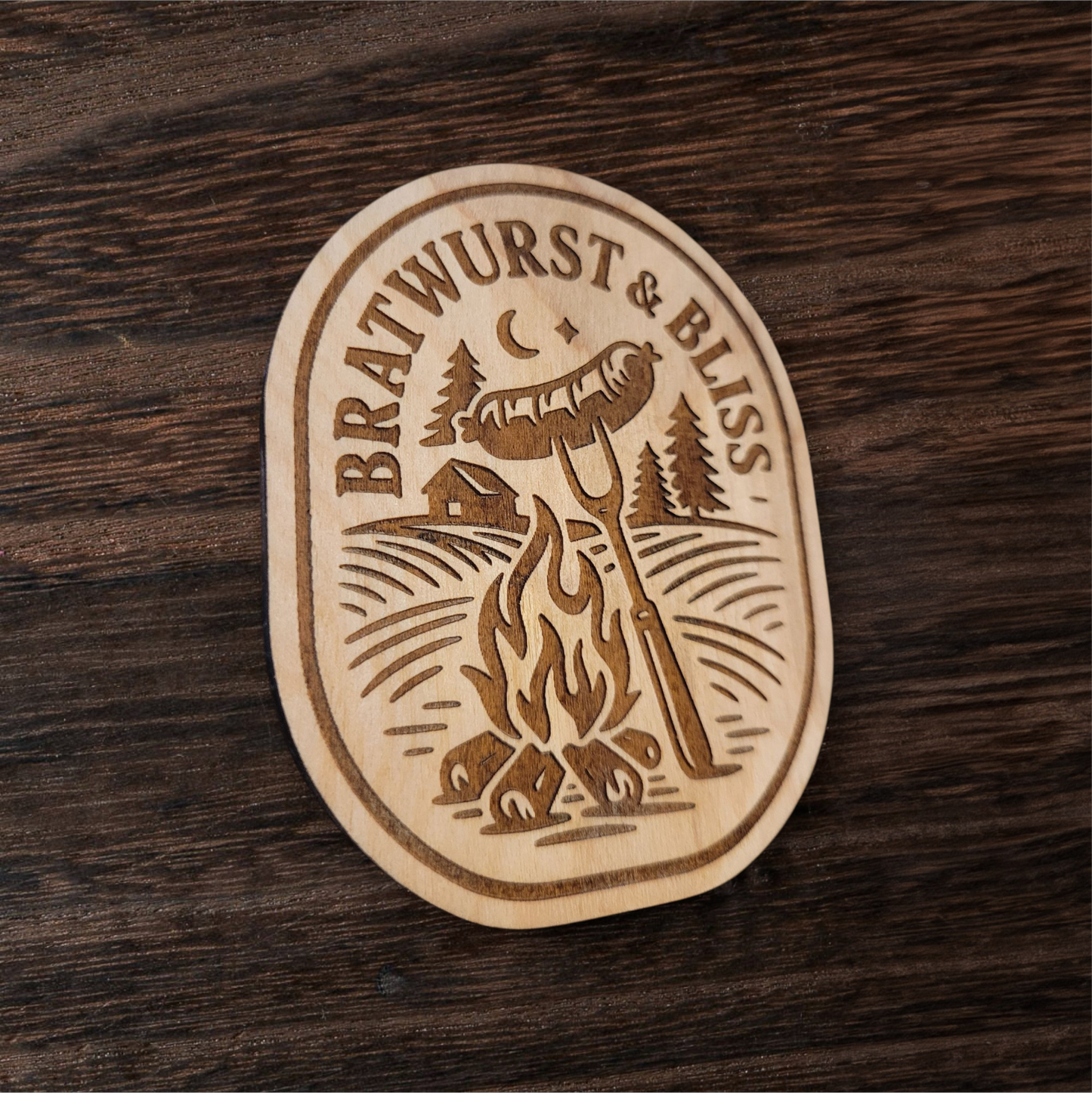 Wood magnet featuring a bratwurst roasting over a campfire with pine trees, cabin, and crescent moon – engraved text reads “Bratwurst & Bliss”