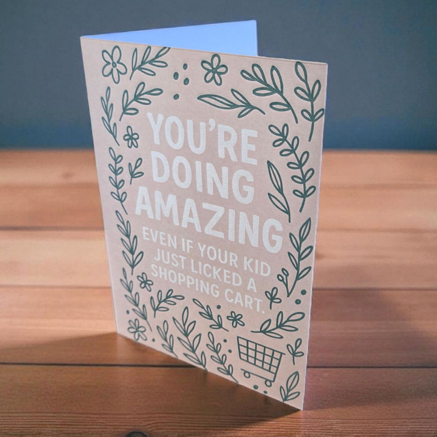 Greeting card reading ‘You’re Doing Amazing Even If Your Kid Just Licked a Shopping Cart’ with green floral border on blush background.