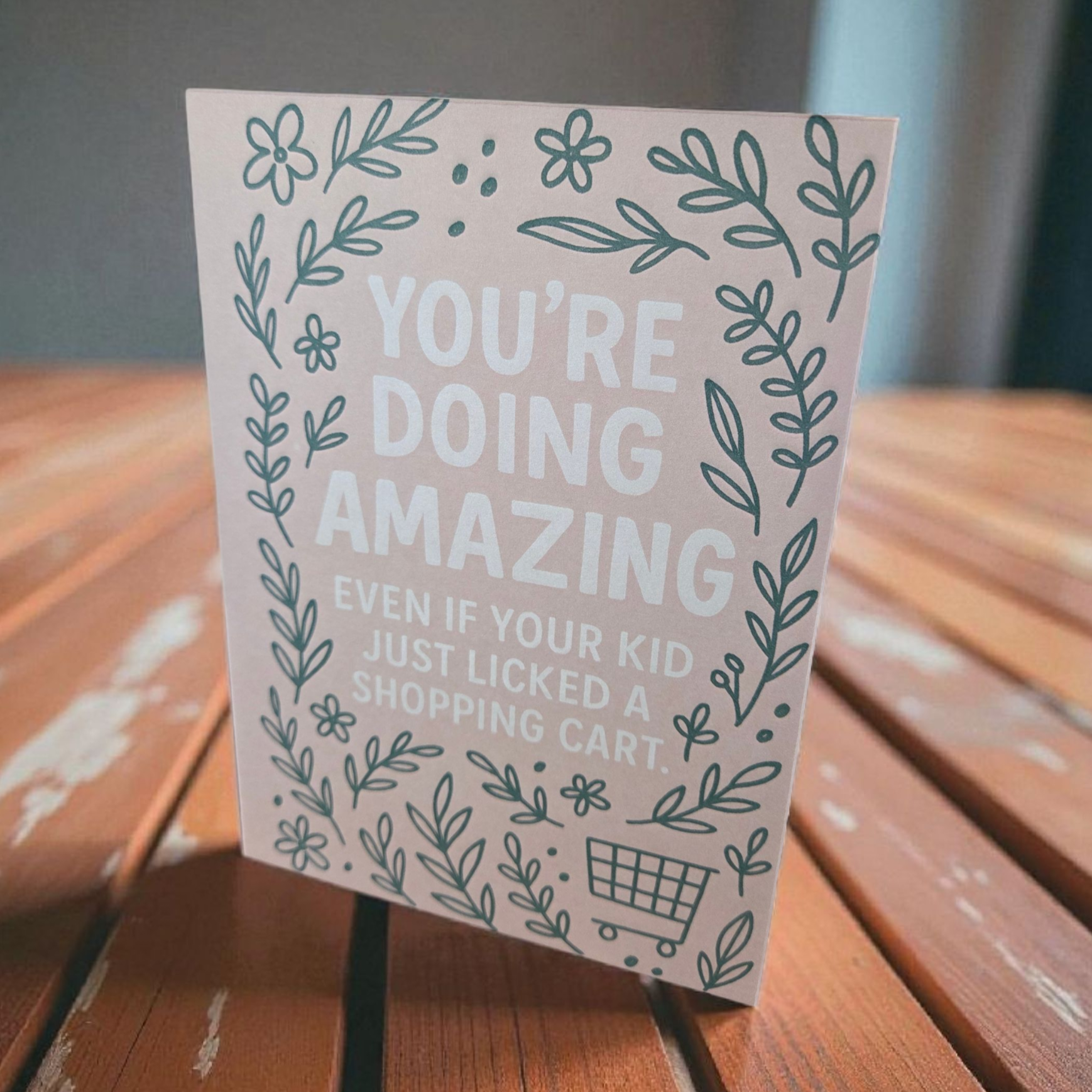 Greeting card reading ‘You’re Doing Amazing Even If Your Kid Just Licked a Shopping Cart’ with green floral border on blush background.