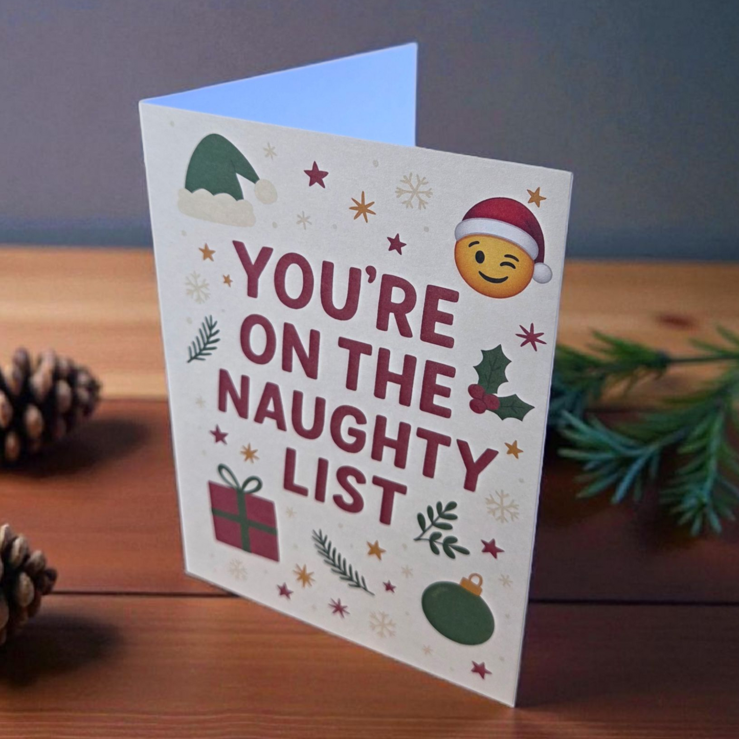 Greeting card reading ‘You’re on the Naughty List’ with Santa hat emoji, holly, ornaments, and stars.