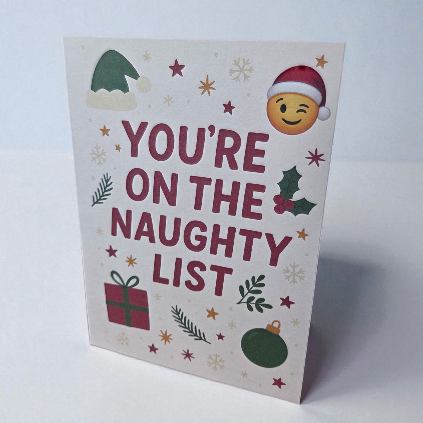 Greeting card reading ‘You’re on the Naughty List’ with Santa hat emoji, holly, ornaments, and stars.