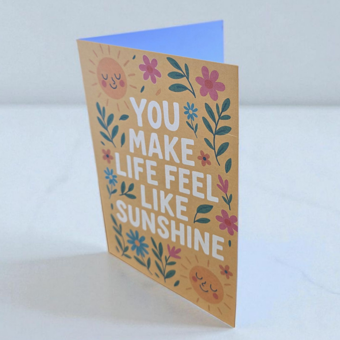 Greeting card reading ‘You Make Life Feel Like Sunshine’ with smiling suns and floral design.