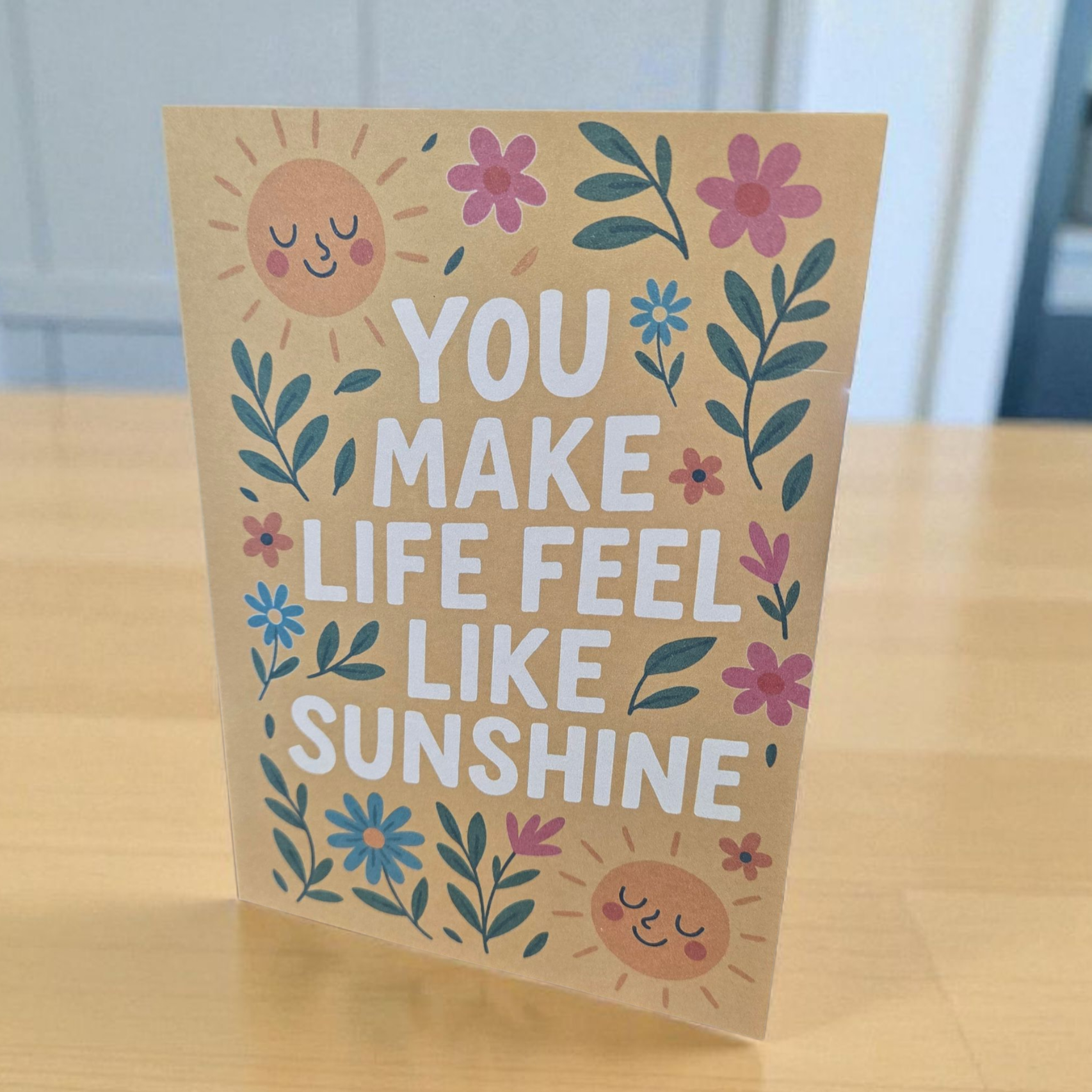 Greeting card reading ‘You Make Life Feel Like Sunshine’ with smiling suns and floral design.