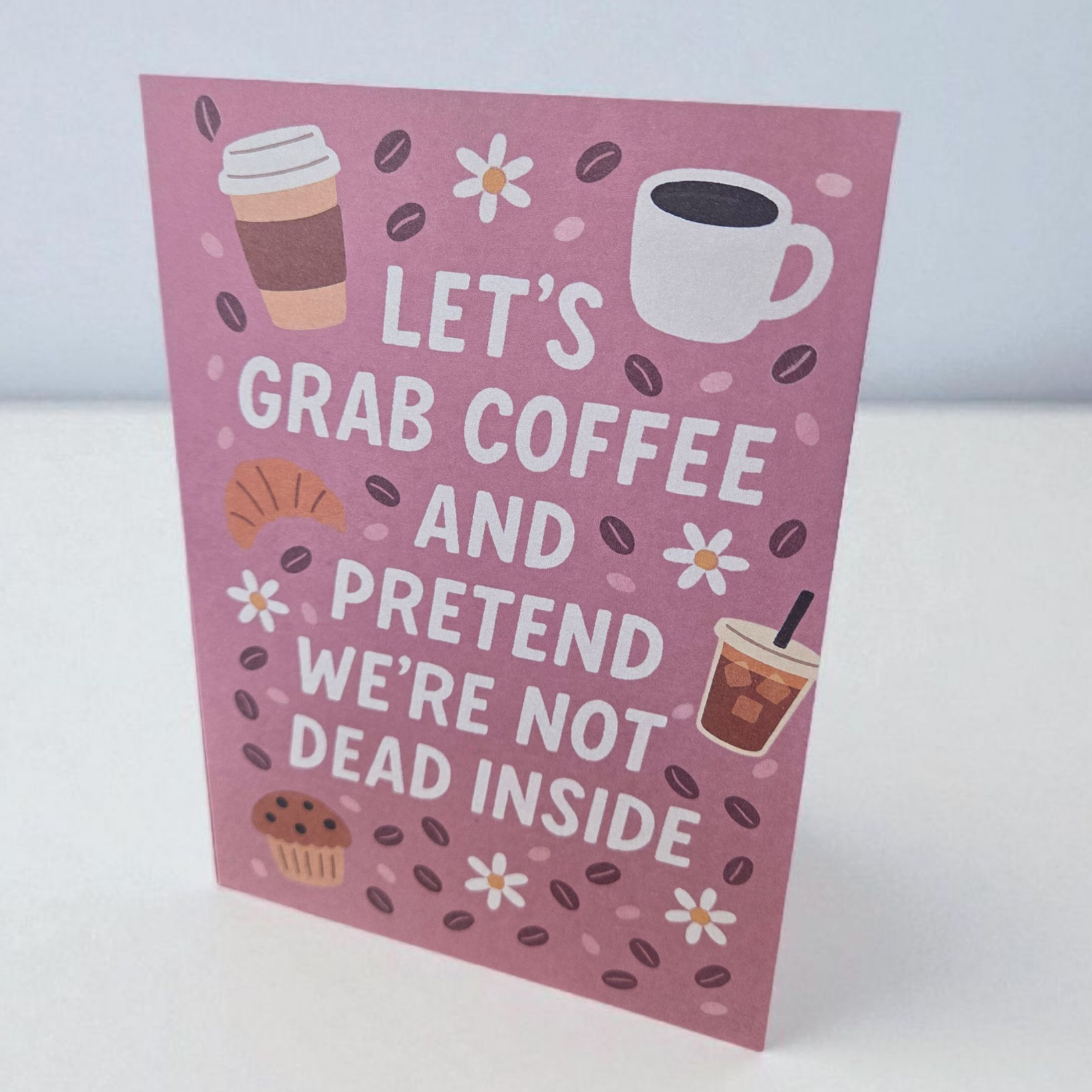 Greeting card reading ‘Let’s Grab Coffee and Pretend We’re Not Dead Inside’ with coffee and pastry illustrations.