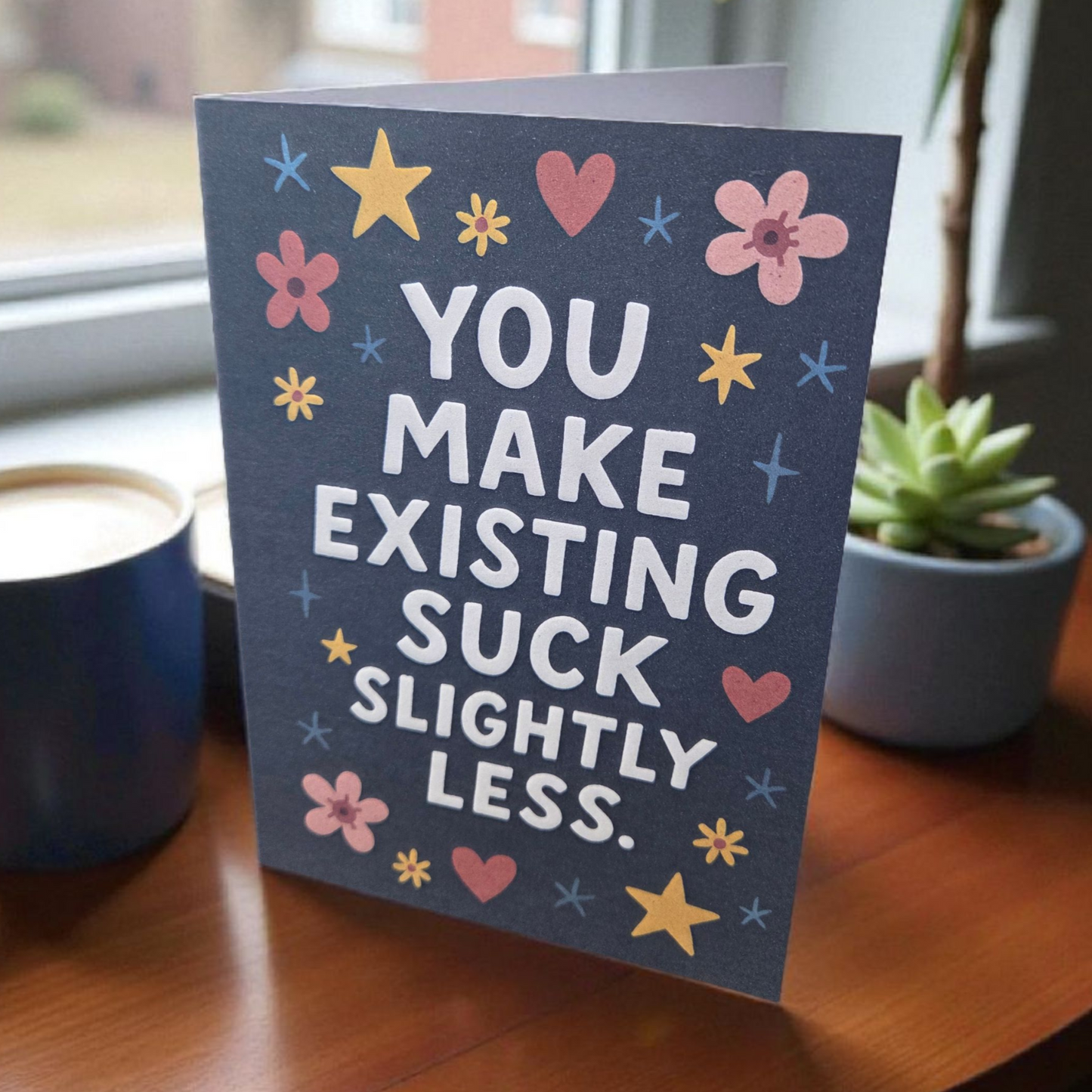 Greeting card reading ‘You Make Existing Suck Slightly Less’ with stars and floral illustrations.