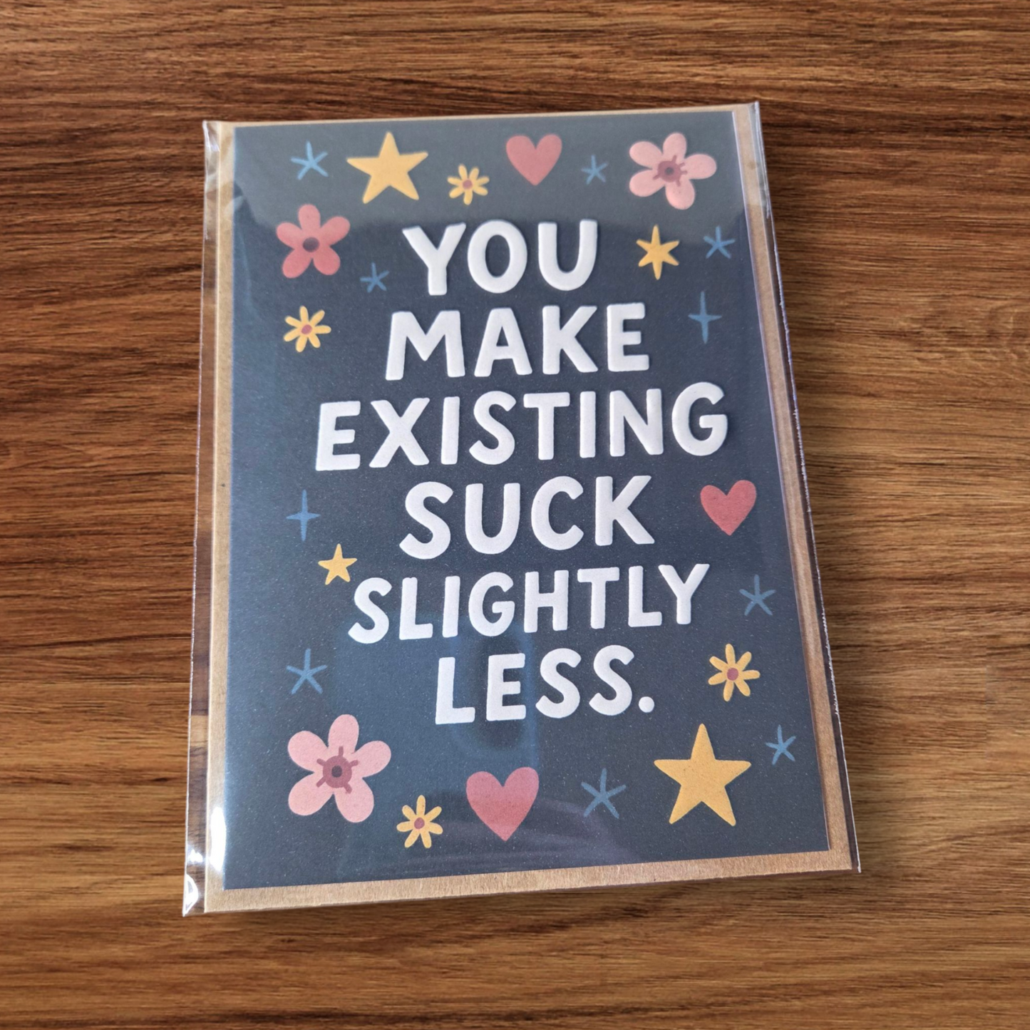 Greeting card reading ‘You Make Existing Suck Slightly Less’ with stars and floral illustrations.