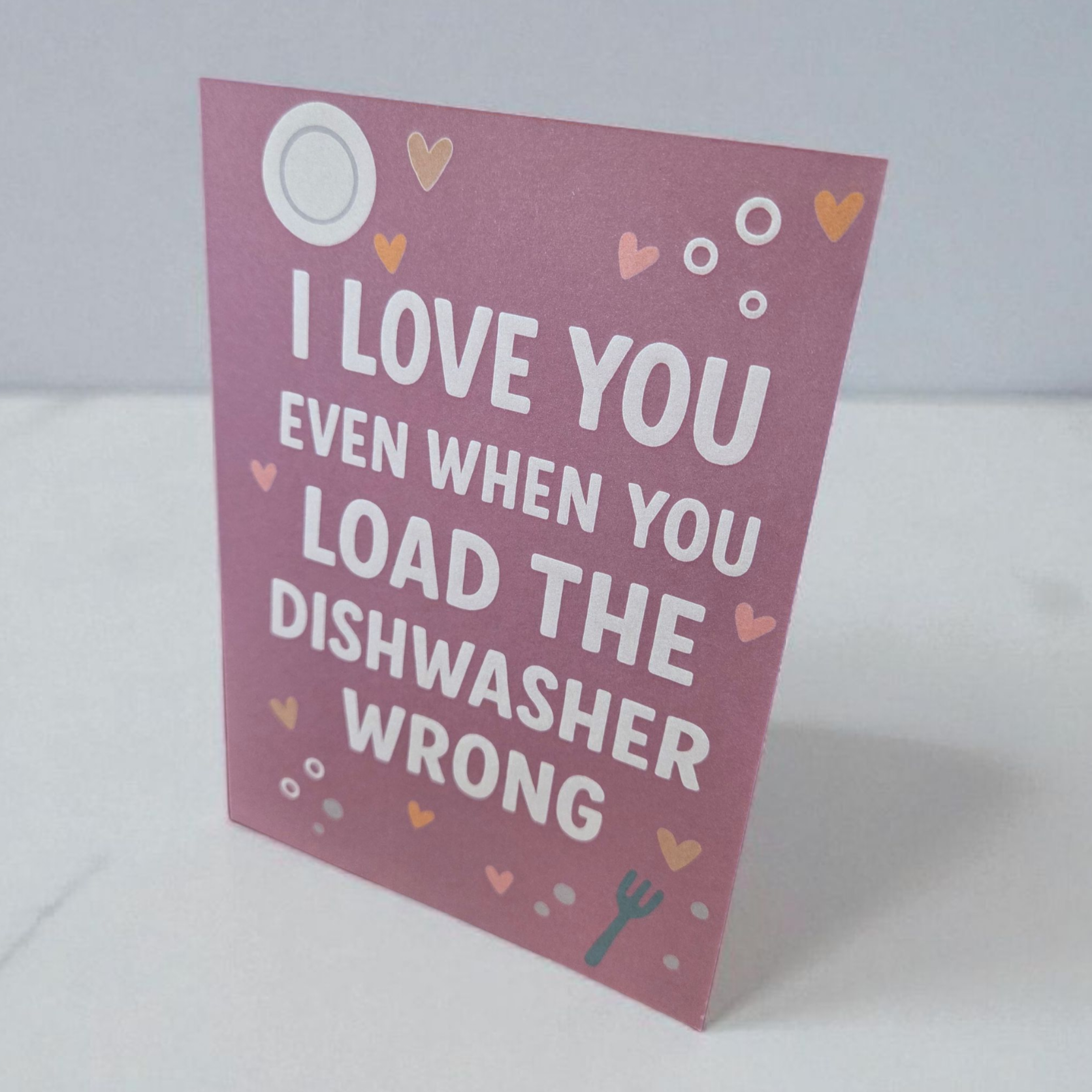 Funny greeting card reading ‘I Love You Even When You Load the Dishwasher Wrong’ on pink background with hearts.