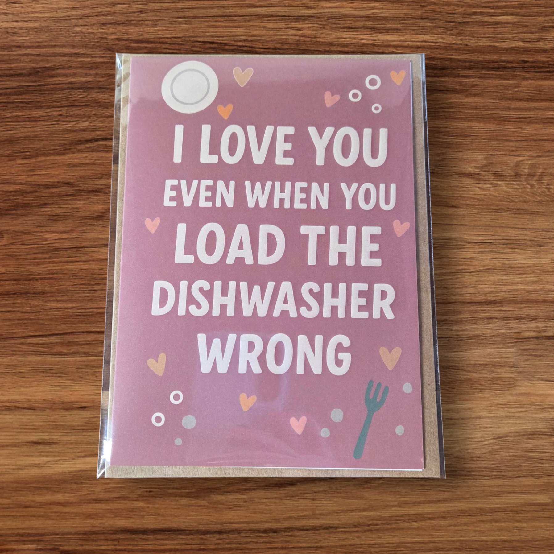 Funny greeting card reading ‘I Love You Even When You Load the Dishwasher Wrong’ on pink background with hearts.