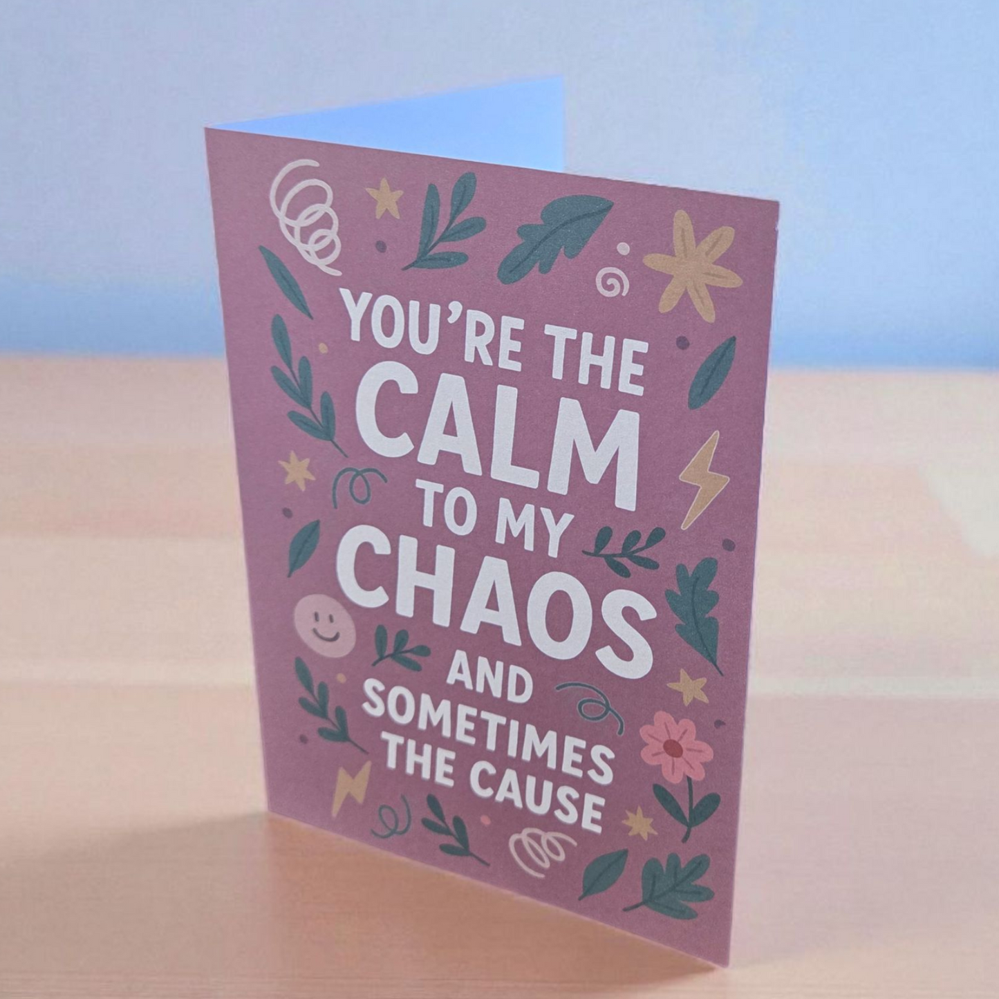 Greeting card reading ‘You’re the Calm to My Chaos and Sometimes the Cause’ on mauve background with floral and lightning illustrations.