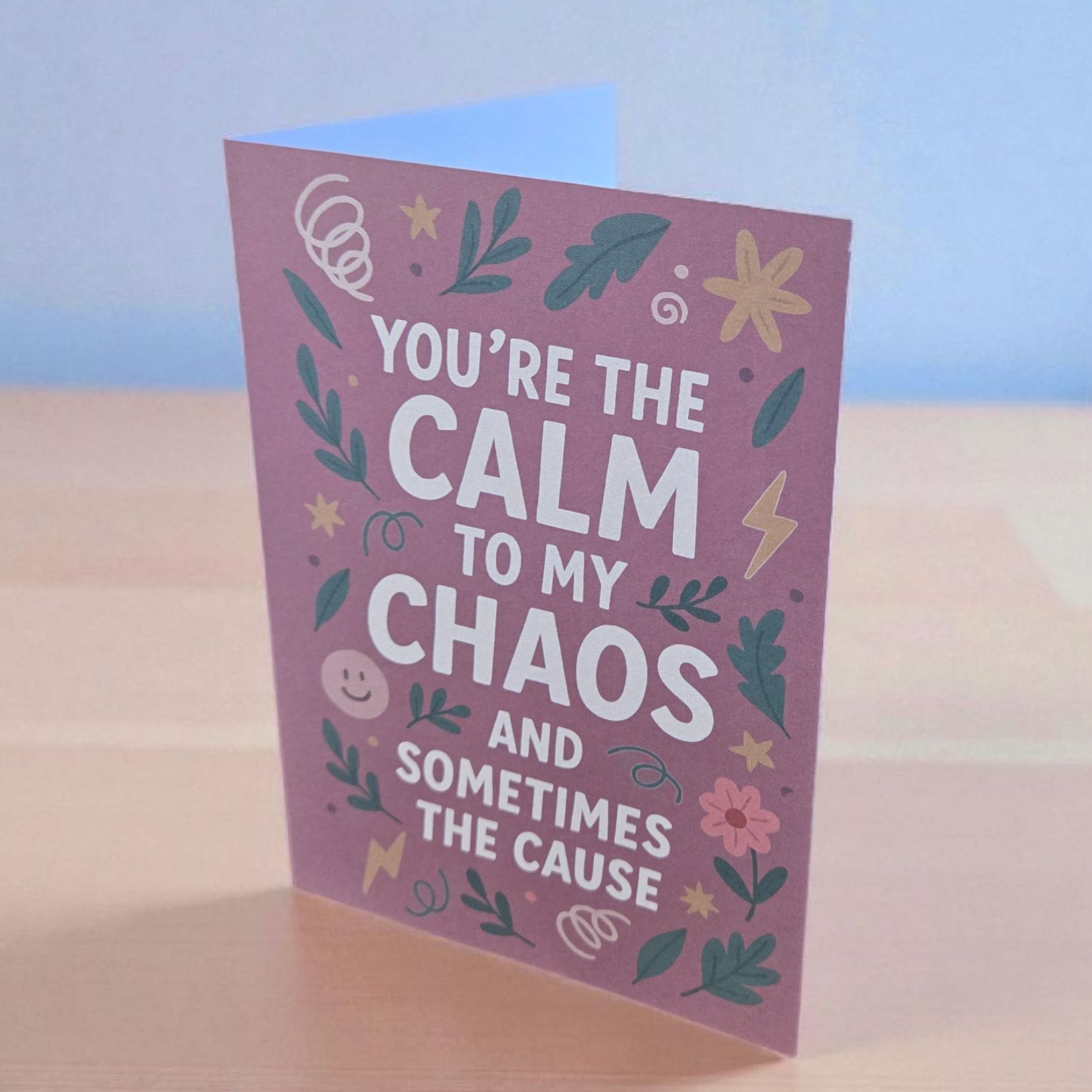 Greeting card reading ‘You’re the Calm to My Chaos and Sometimes the Cause’ on mauve background with floral and lightning illustrations.