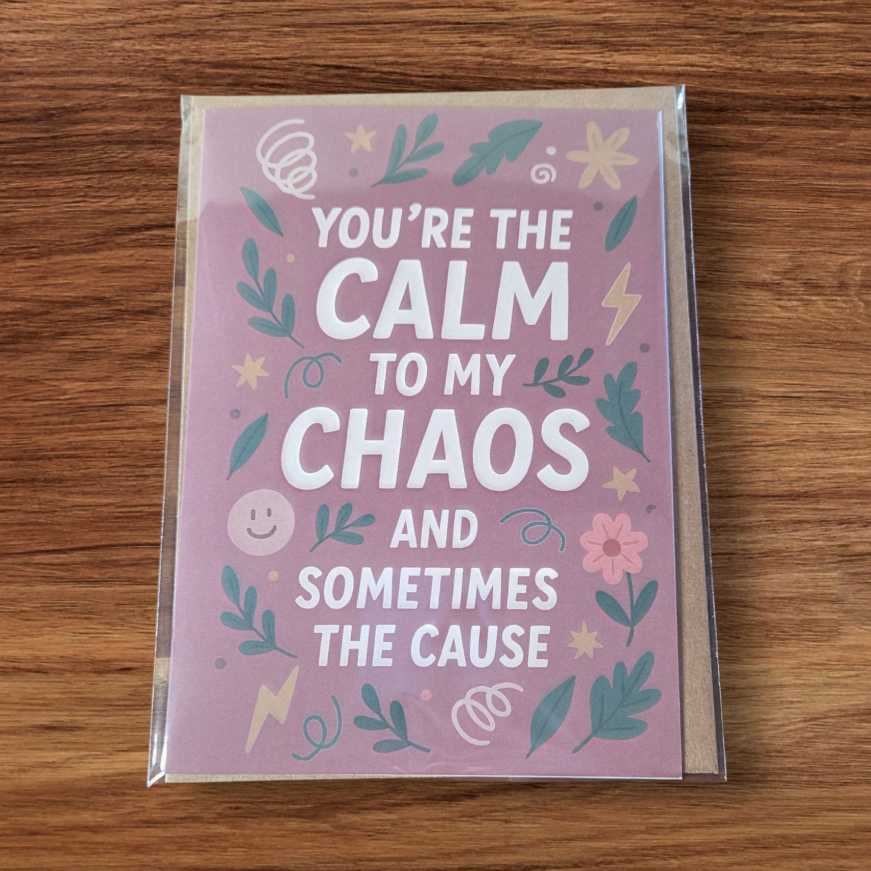 Greeting card reading ‘You’re the Calm to My Chaos and Sometimes the Cause’ on mauve background with floral and lightning illustrations.