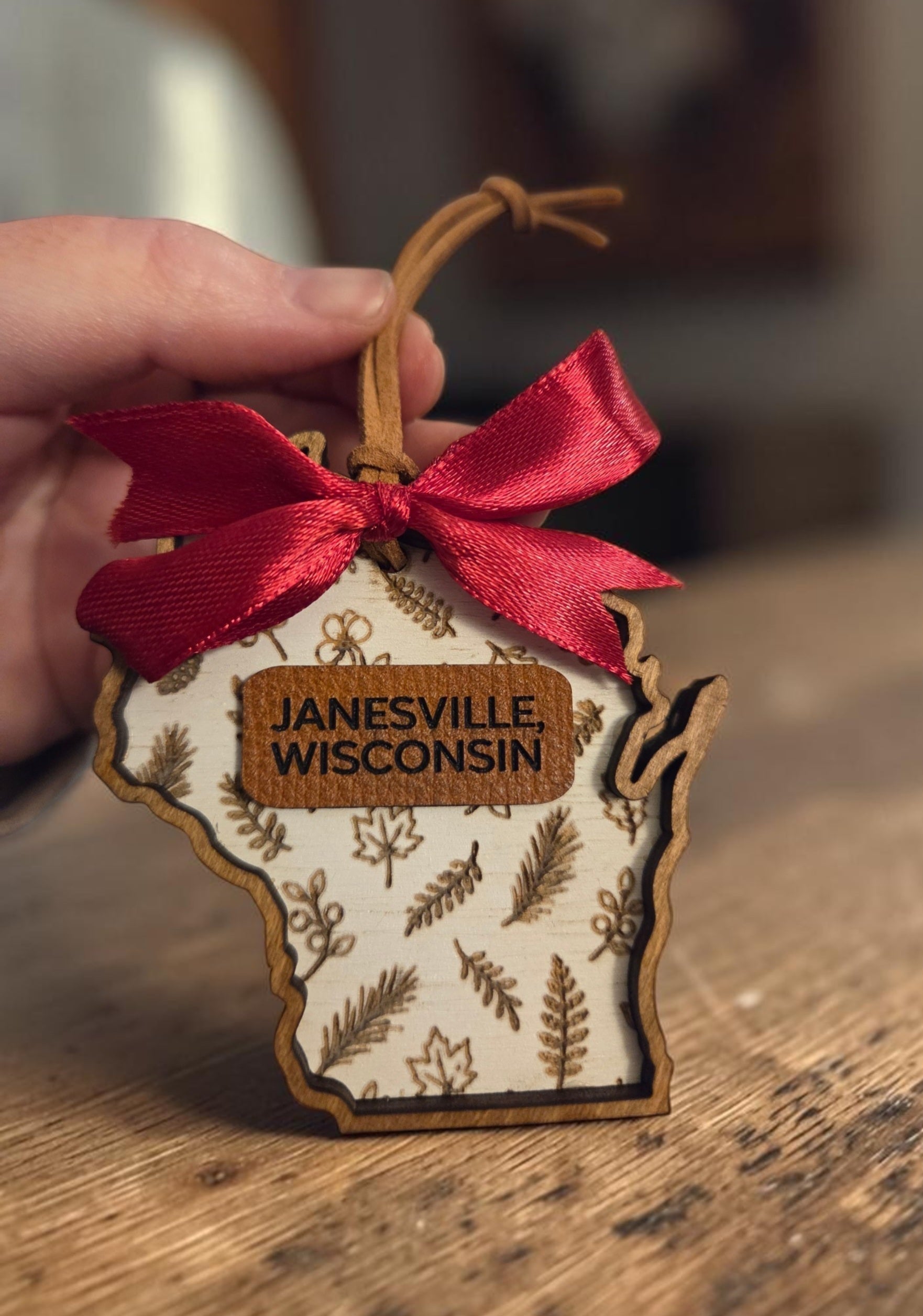 Handcrafted Wisconsin-shaped botanical wood ornament personalized with Janesville, Wisconsin on a leather accent and finished with a wine-red bow and leather hanger.