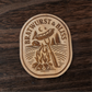 Wood magnet featuring a bratwurst roasting over a campfire with pine trees, cabin, and crescent moon – engraved text reads “Bratwurst & Bliss”