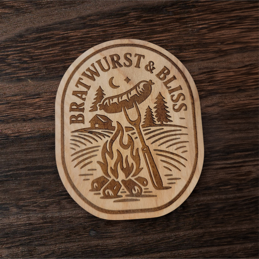 Wood magnet featuring a bratwurst roasting over a campfire with pine trees, cabin, and crescent moon – engraved text reads “Bratwurst & Bliss”