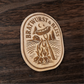 Wood magnet featuring a bratwurst roasting over a campfire with pine trees, cabin, and crescent moon – engraved text reads “Bratwurst & Bliss”