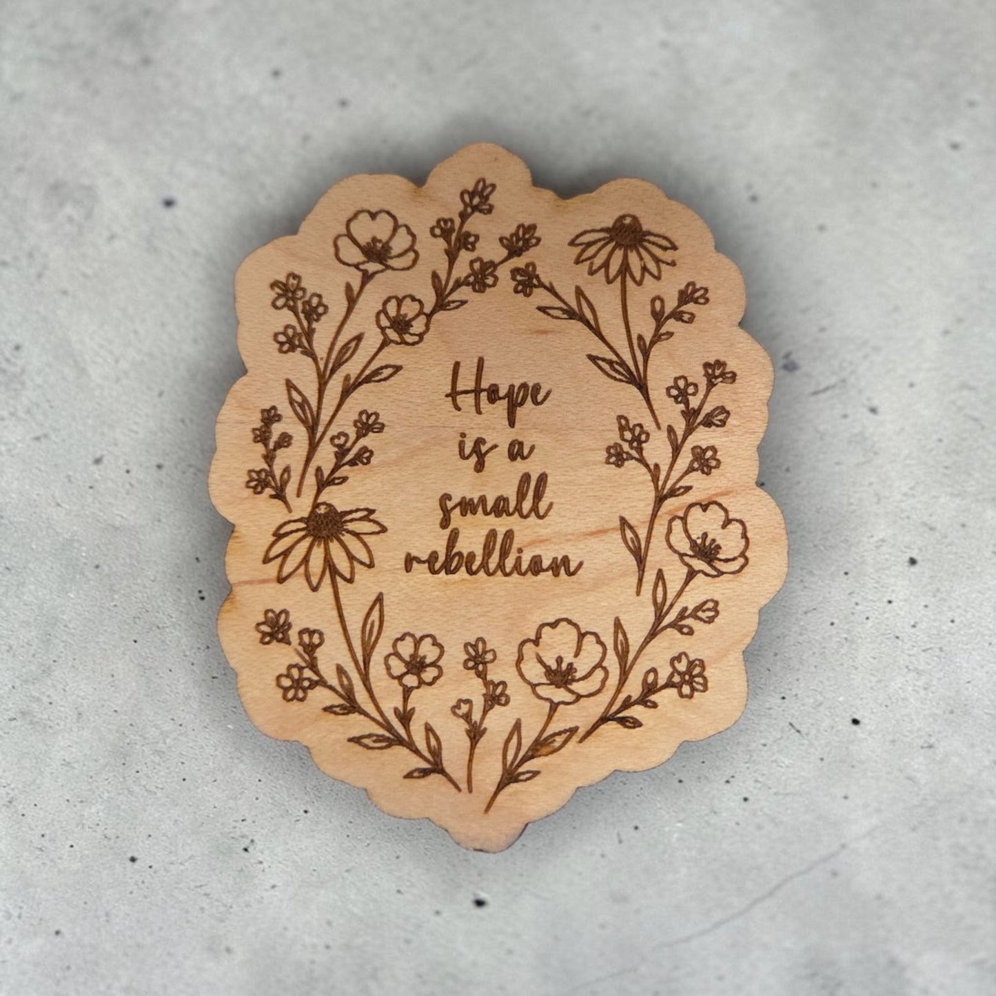 Handcrafted natural wood magnet engraved with the phrase “Hope is a small rebellion” surrounded by delicate wildflowers on a neutral gray background.