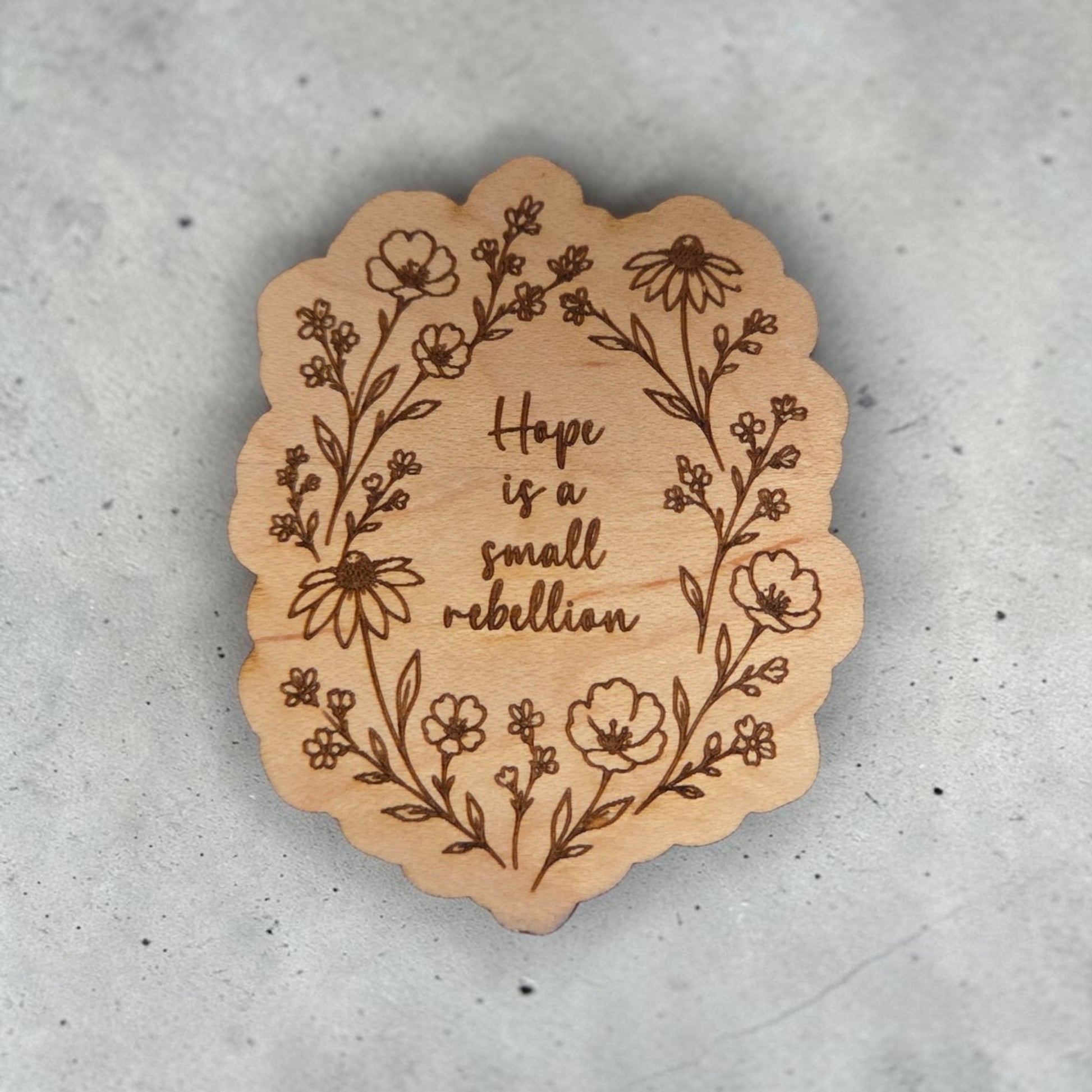 Handcrafted natural wood magnet engraved with the phrase “Hope is a small rebellion” surrounded by delicate wildflowers on a neutral gray background.