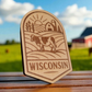 Laser-engraved wood magnet with a cow, barn, cornfield, and rising sun in a scenic Wisconsin farm badge design.