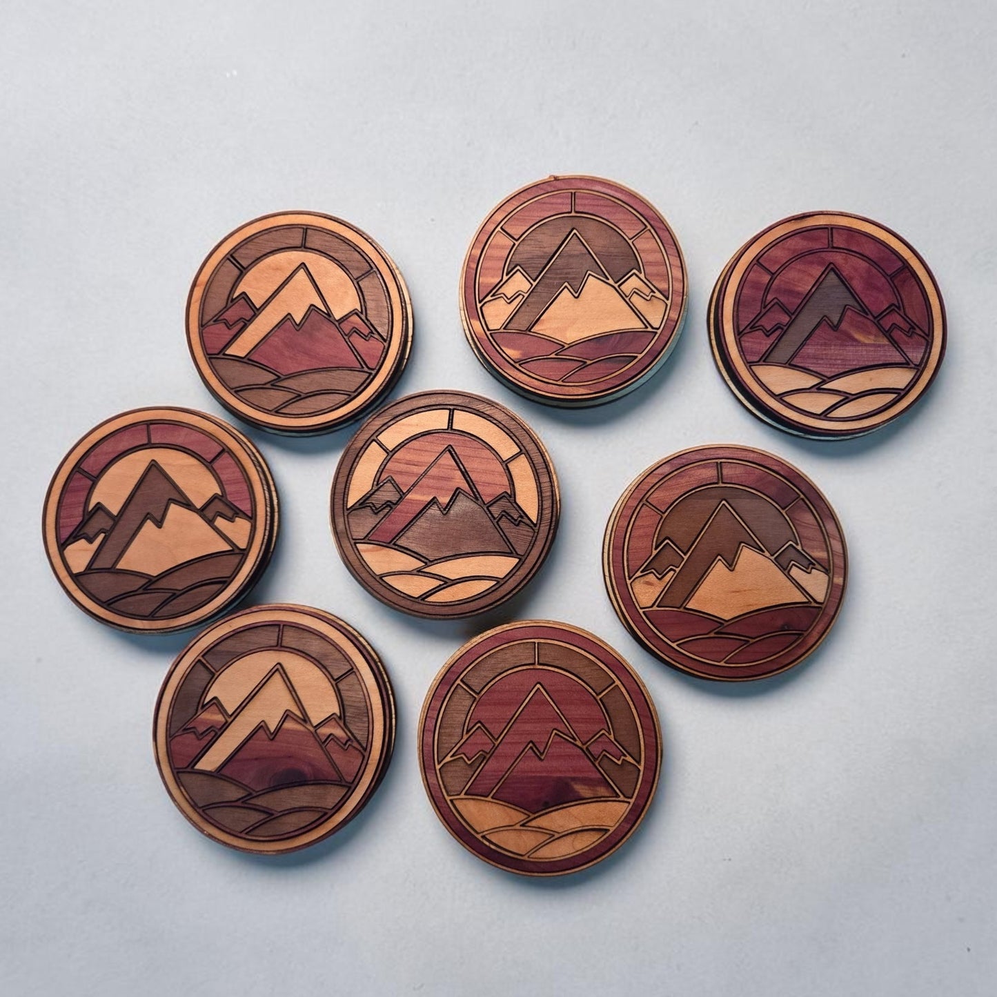 Example assortment of handcrafted round mountain magnets made from layered cedar, walnut, and maple wood. Each magnet features a unique combination of natural wood tones and mountain details. Listing includes one randomly selected magnet; exact design pictured is not guaranteed.