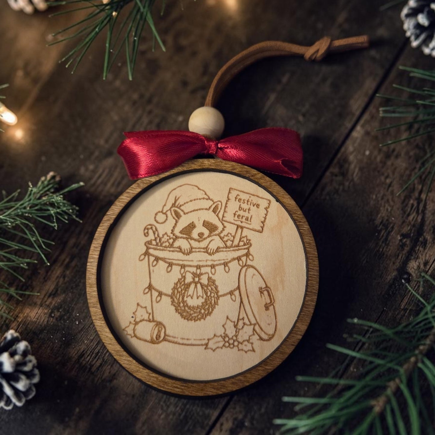 Handcrafted layered wood Christmas ornament featuring an engraved raccoon in a festively decorated trash can holding a “festive but feral” sign, finished with a wine-red bow, wood bead, and leather hanging loop.
